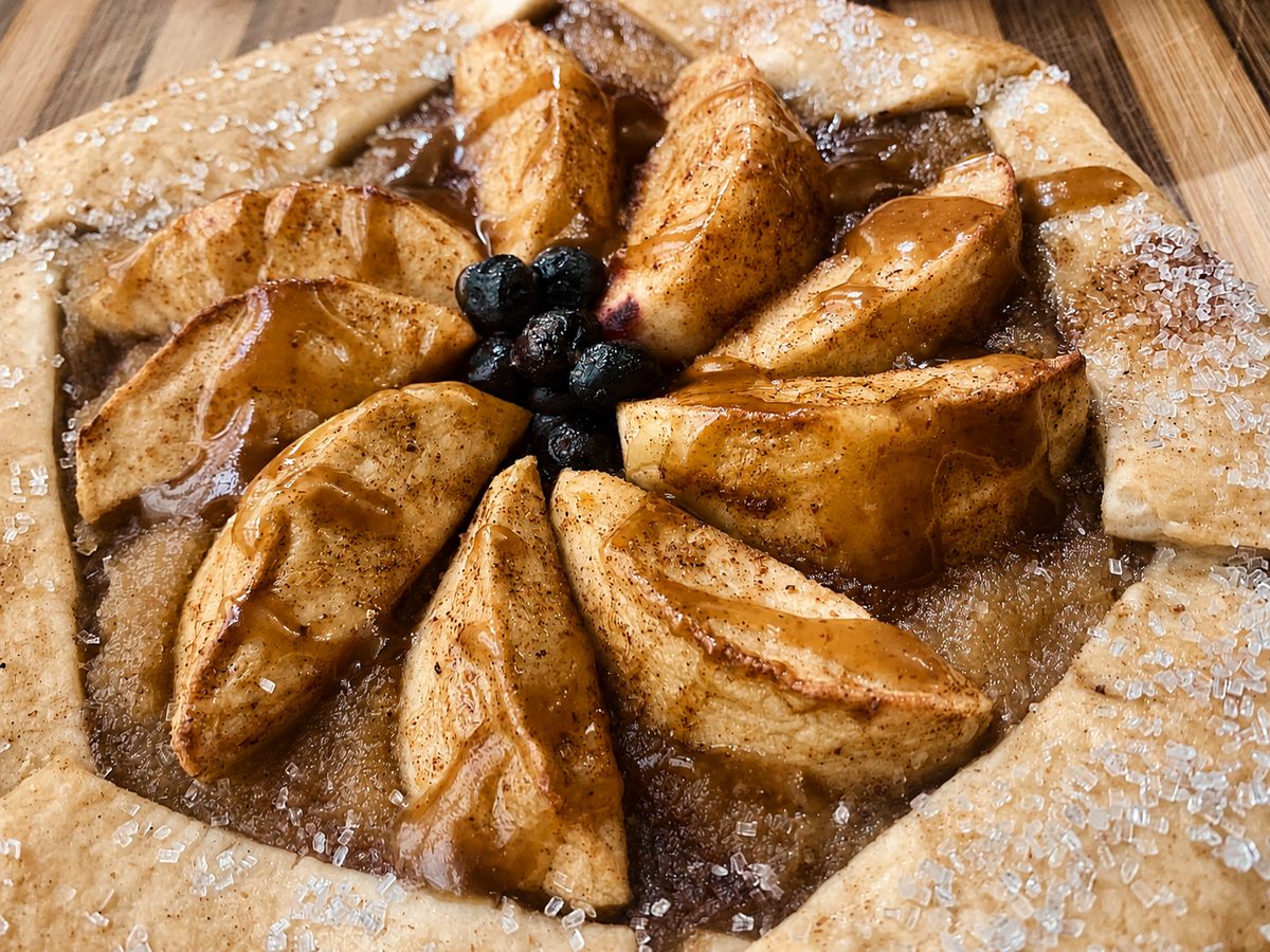 A rustic vegan apple galette with blueberries at the centre, cinnamon, a caramel drizzle and a sugared crust, on a wooden board.
