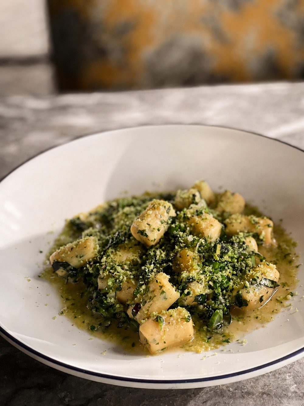 Vegan potato gnocchi with wilted greens in an herb sauce, finished with a green herb crumb, in a white bowl with a navy rim.
