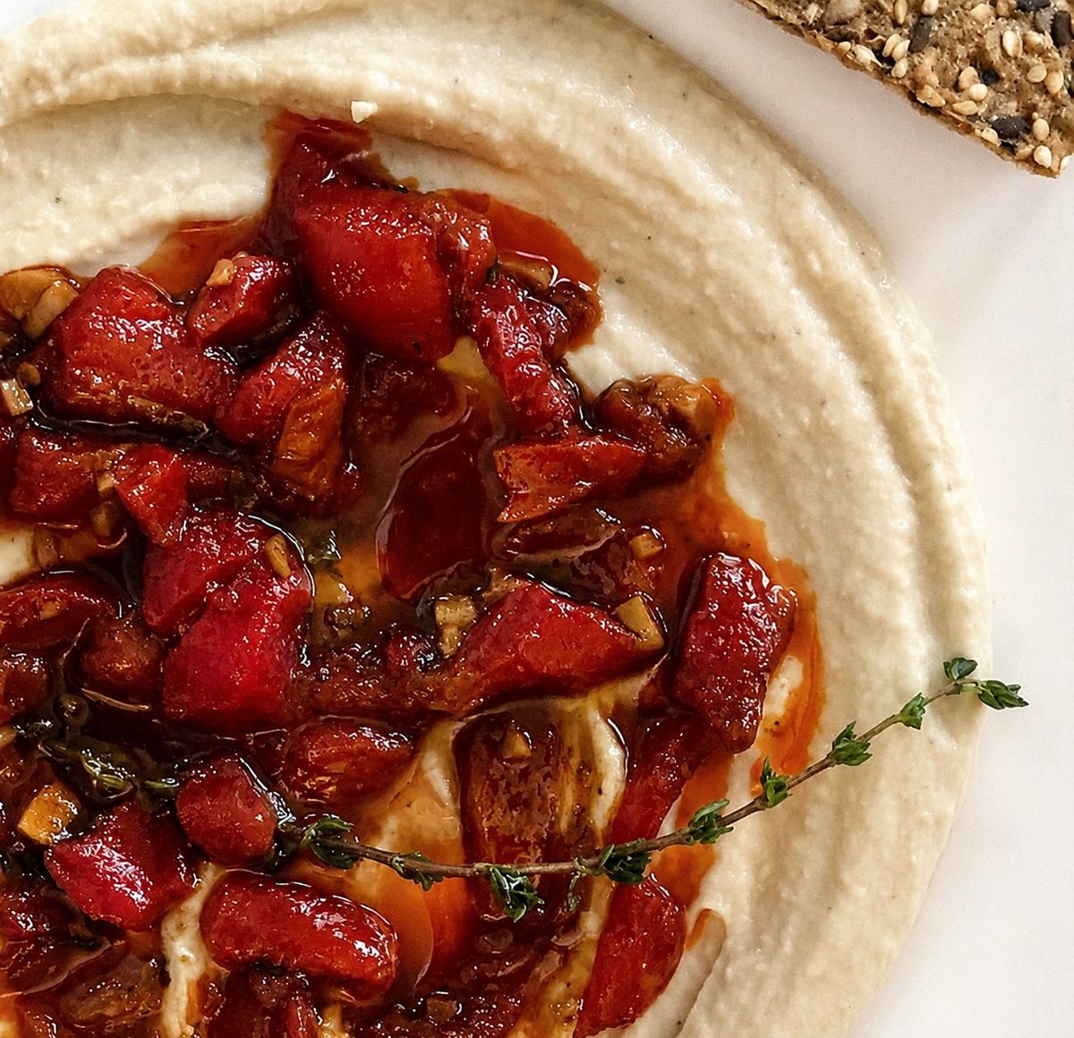 Vegan whipped white bean purée swirled on a white plate, topped with roasted red peppers, garlic, olive oil and a sprig of thyme, with a seeded cracker at the edge.