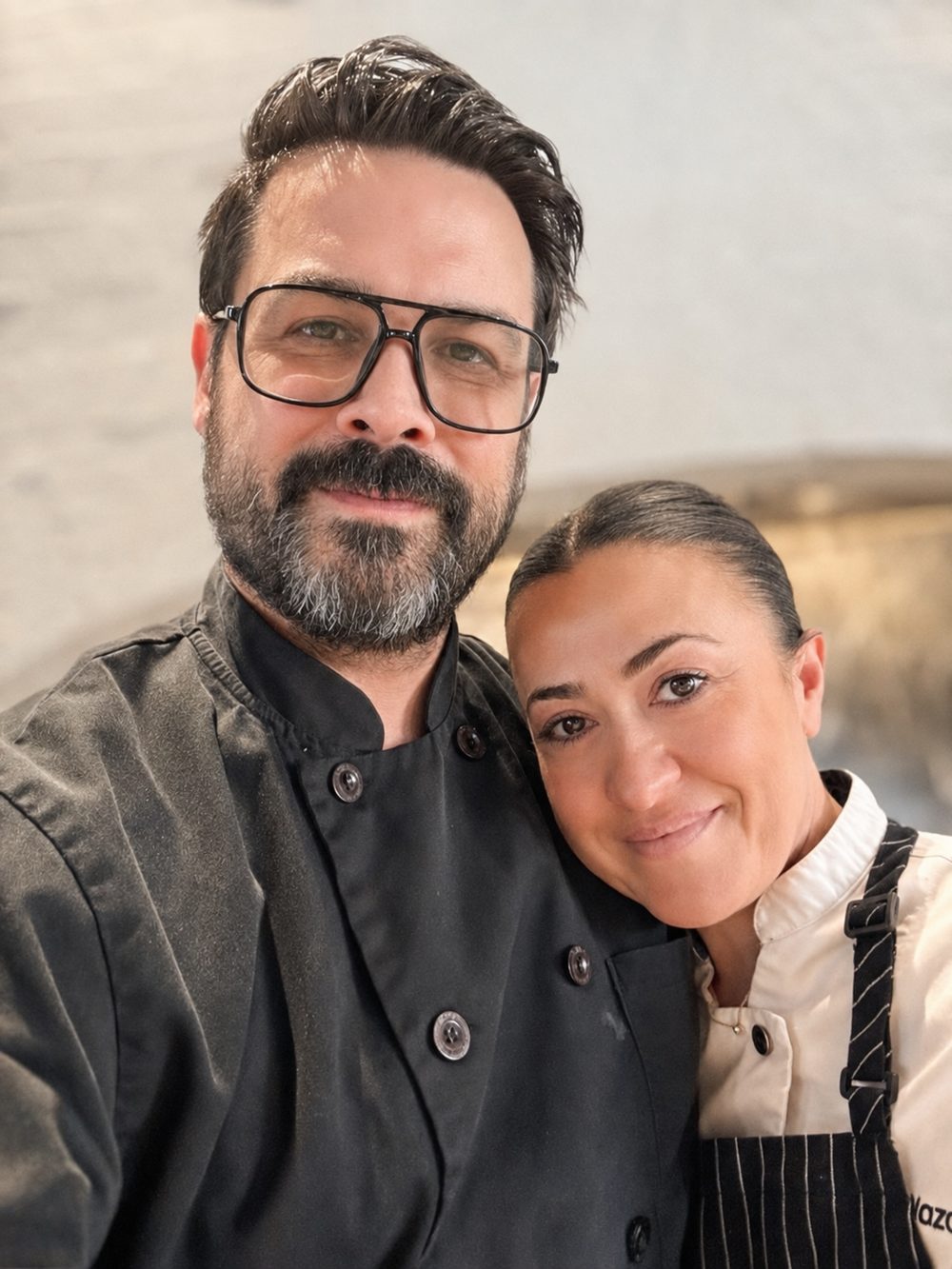 Chef Jesse Brune and Chef Manifa Nazarian in chef jackets, heads together and smiling, in a kitchen.