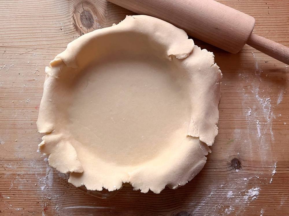 A vegan pastry crust pressed into a pie dish on a floured wooden bench, rolling pin alongside.